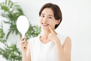 woman with short brown hair in a white shirt holding a mirror and pointing to her smile