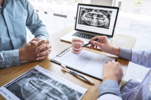 Two men sitting at a desk that has a toothbrush, paperwork, model teeth, and dental X-rays as if discussing